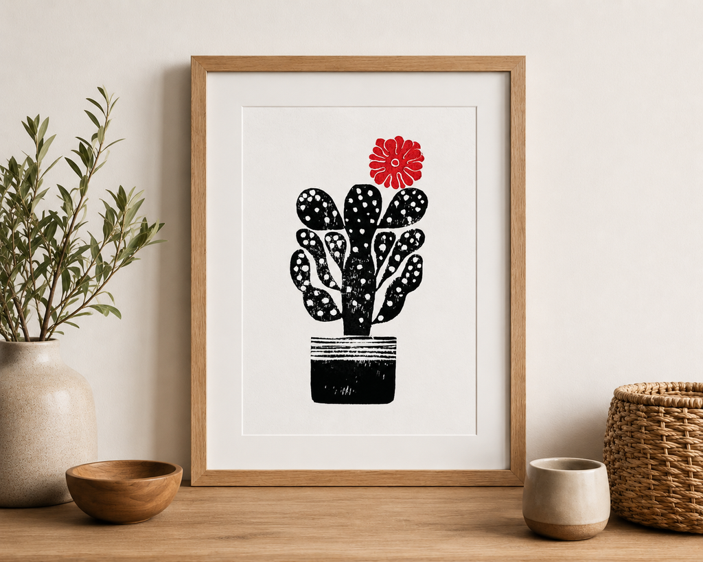 handmade linocut print of a cactus with a flower in black and red ink, A4 size, signed artwork on a sideboard in a room with plants and scandi decor