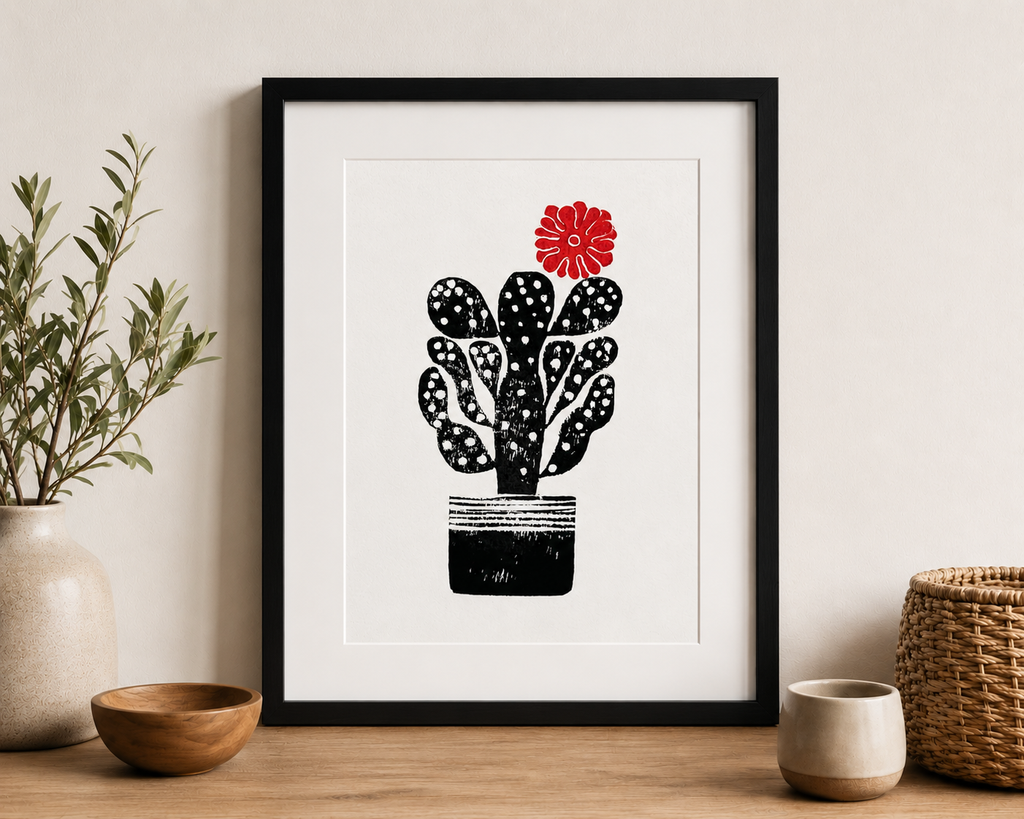 handmade linocut print of a cactus with a flower in black and red ink, A4 size, signed artwork on a sideboard in a room with plants and scandi style decor
