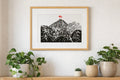 Handmade linocut mountain and love heart print, framed, black and red ink on white textured paper, A4 size, set on a wall above a wooden shelf with plants.