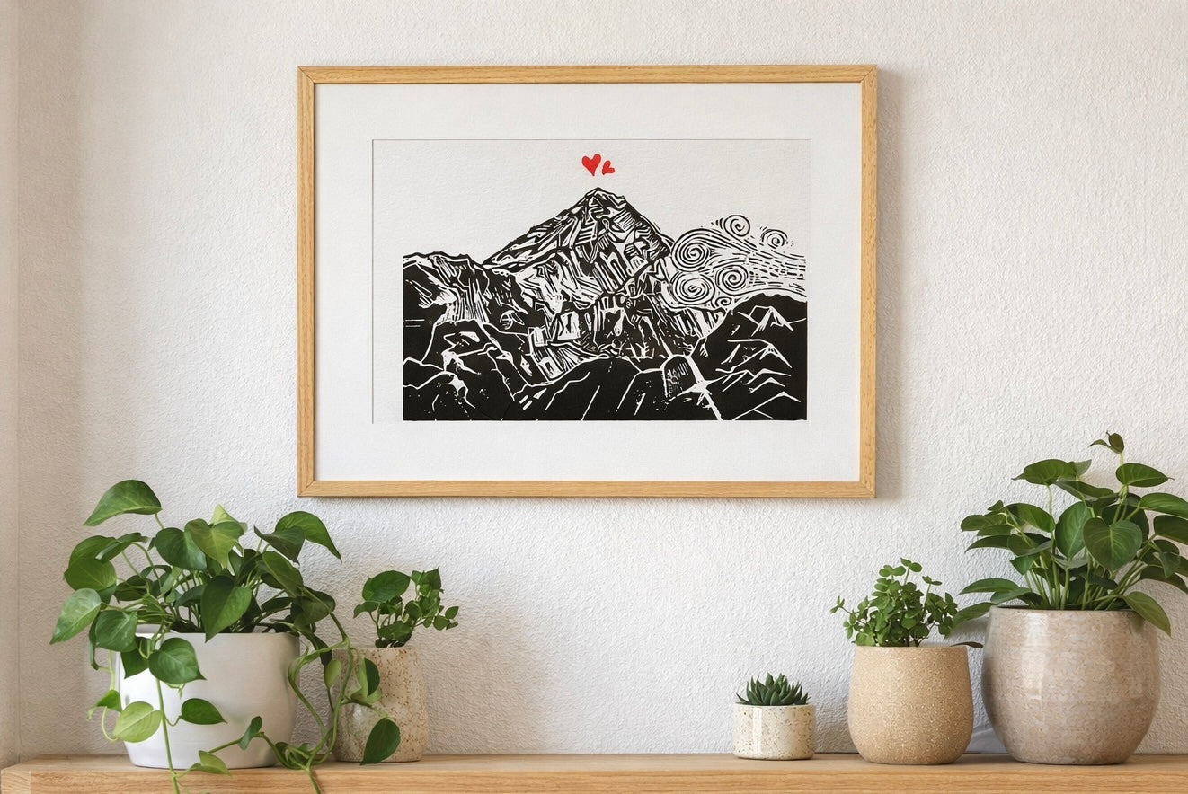 Handmade linocut mountain and love heart print, framed, black and red ink on white textured paper, A4 size, set on a wall above a wooden shelf with plants.
