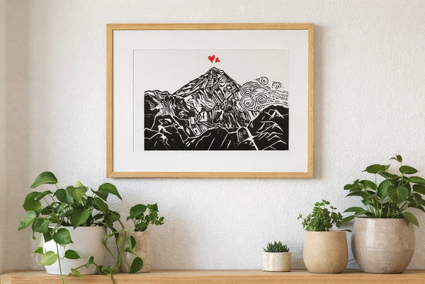 Handmade linocut mountain and love heart print, framed, black and red ink on white textured paper, A4 size, set on a wall above a wooden shelf with plants.
