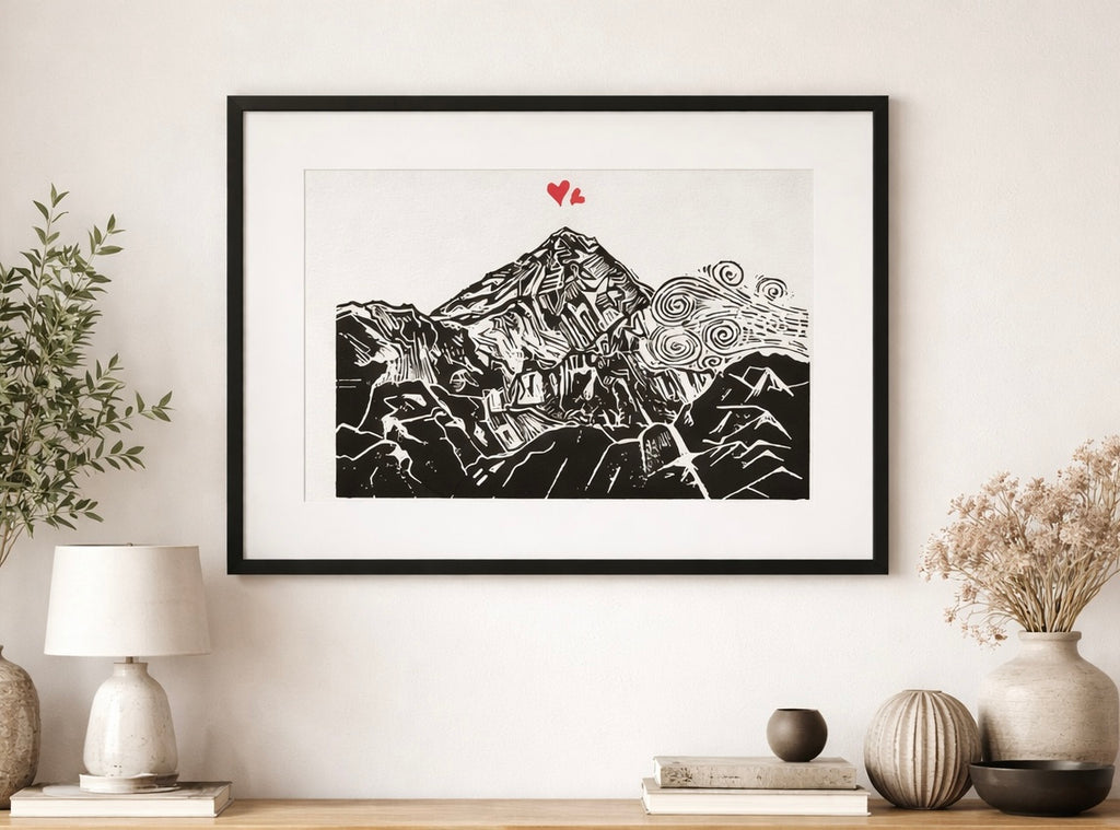 Handmade linocut mountain and love heart print, framed, black and red ink on white textured paper, A4 size, set on a wall above a wooden shelf with plants and scandi style decor.