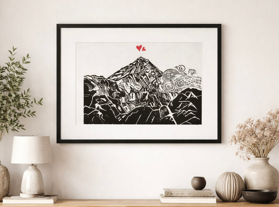 Handmade linocut mountain and love heart print, framed, black and red ink on white textured paper, A4 size, set on a wall above a wooden shelf with plants and scandi style decor.