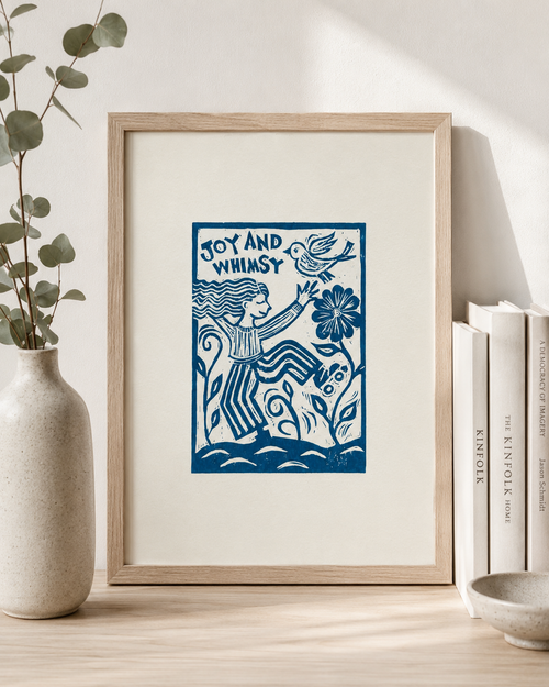 Framed artwork with blue design and text on a white wall, next to a vase and books.