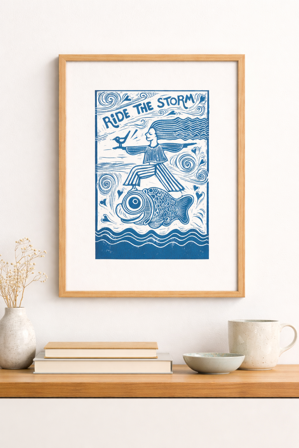 Framed artwork with a blue design of a person riding a fish on a white wall above a wooden shelf.