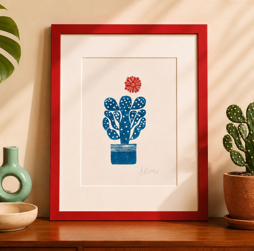 handmade linocut print of a cactus with a flower in turquoise and red ink, A4 size, signed artwork on a sideboard in a room with plants and decor