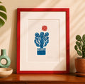 handmade linocut print of a cactus with a flower in turquoise and red ink, A4 size, signed artwork on a sideboard in a room with plants and decor